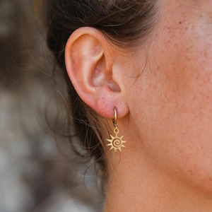 May include: A gold-colored sunburst charm earring with a spiral center is shown. The earring is attached to a small hoop that is attached to the ear. The earring is hanging from the ear.