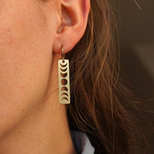 May include: Gold dangle earrings with a rectangular charm featuring a cutout design of the moon phases.