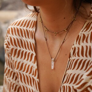 May include: A layered necklace with a crystal pendant. The top necklace is made of brown cord with small gold beads. The bottom necklace is made of brown cord with a clear crystal pendant.