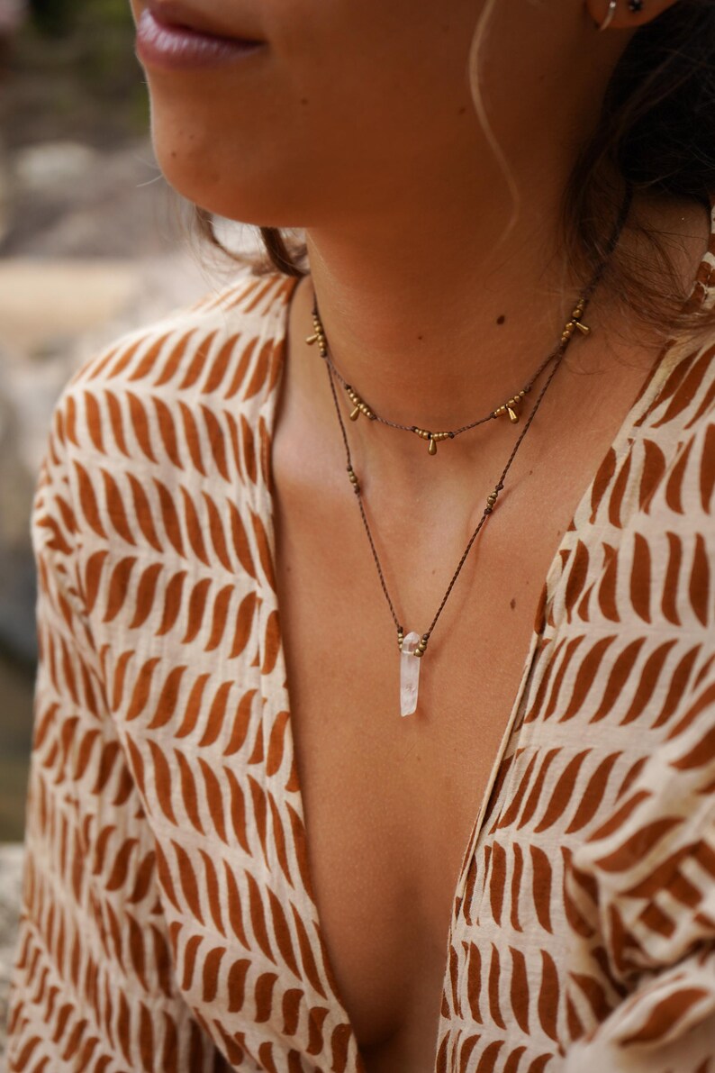 May include: A layered necklace with a crystal pendant. The top necklace is made of brown cord with small gold beads. The bottom necklace is made of brown cord with a clear crystal pendant.