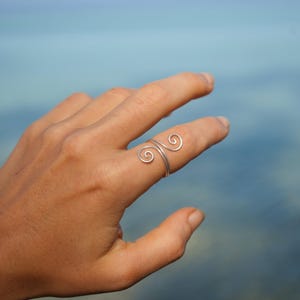 Könnte beinhalten: Ein silberner Ring mit einem Doppelspiralen-Design. Der Ring wird am Mittelfinger einer Hand getragen.
