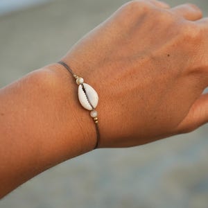 Può includere: Braccialetto minimalista con un pendente a conchiglia bianca naturale. La conchiglia è infilata su un sottile cordoncino marrone scuro, impreziosito da piccole perline color oro. Il braccialetto ha un design semplice, ispirato alla spiaggia.