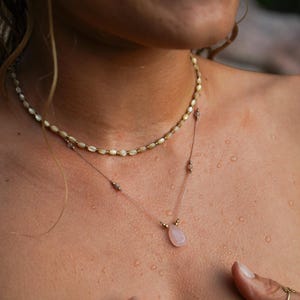 Può includere: Una collana a due fili con un ciondolo di pietra rosa. La collana superiore è composta da piccole perle ovali iridescenti. La collana inferiore è una catena delicata con un ciondolo di pietra rosa.