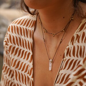 May include: A woman wearing a brown and white patterned shirt with a layered necklace featuring a clear quartz crystal pendant and gold beads.