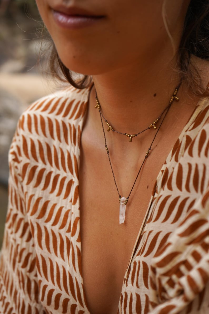 May include: A woman wearing a brown and white patterned shirt with a layered necklace featuring a clear quartz crystal pendant and gold beads.