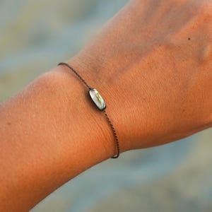 Könnte beinhalten: Ein filigranes Armband mit einem kleinen, ovalen, irisierenden Anhänger. Der Anhänger zeigt eine Mischung aus Farben, darunter Grün, Weiß und Gold. Das Armband besteht aus einer dünnen, dunklen Kette.