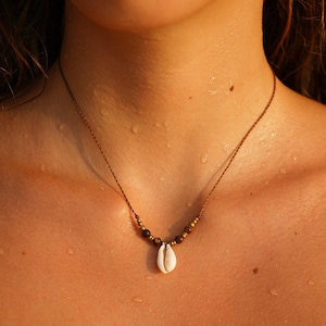 Op de afbeelding: Een delicate ketting met een natuurlijke schelpenhanger. De ketting heeft een bruin koord met kleine gouden en bruine kralen. De schelp is wit met een glad oppervlak. De ketting wordt op de huid gedragen.