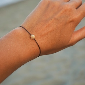 Könnte beinhalten: Ein minimalistisches Armband mit einer kleinen, runden, hellfarbigen Perle in der Mitte eines dunkelbraunen Bandes. Das Armband ist schlicht und elegant, für den Alltag geeignet. Der Hintergrund ist unscharf, was auf eine Umgebung im Freien hindeutet.