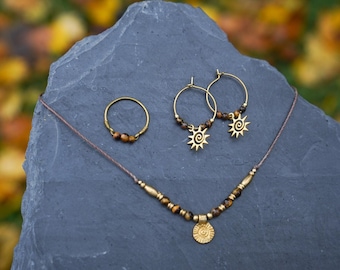 May include: A set of gold jewelry featuring a necklace with a sun pendant, a ring, and a pair of earrings. The necklace is made with brown tiger's eye beads and a gold sun pendant. The ring is a simple gold band with a tiger's eye stone. The earrings are gold hoops with a sun pendant on each.