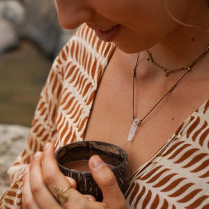 May include: A person holding a brown coconut shell cup with a brown liquid inside. The person is wearing a white and brown patterned shirt and a necklace with a clear crystal pendant.
