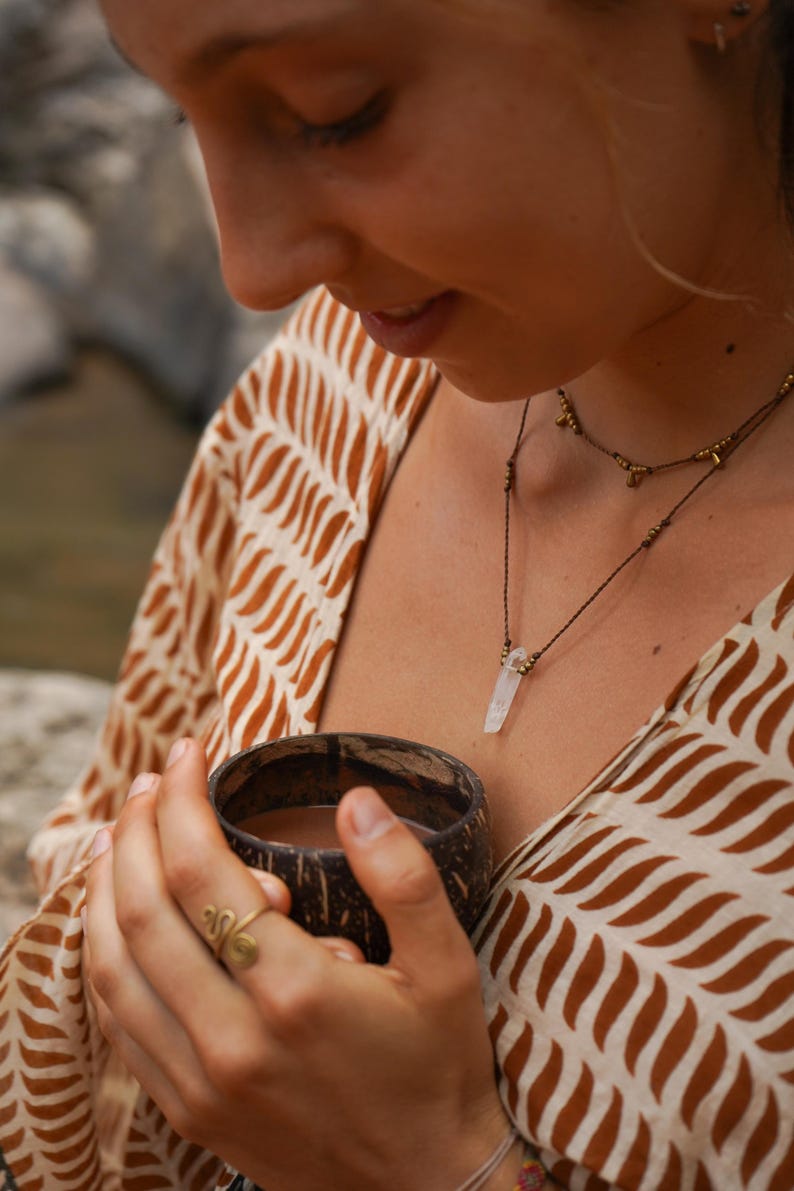 May include: A person holding a brown coconut shell cup with a brown liquid inside. The person is wearing a white and brown patterned shirt and a necklace with a clear crystal pendant.