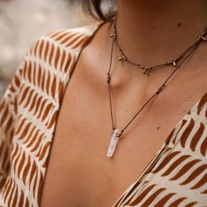 May include: A delicate necklace with a clear quartz crystal pendant and gold accents. The necklace is made with brown cord and small gold beads.