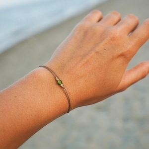 Peut inclure: Un bracelet délicat avec un fin cordon marron, une perle vert olive et deux petites perles dorées. Le bracelet est porté sur un poignet, avec un arrière-plan flou de plage et d'océan.