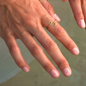 Könnte beinhalten: Ein goldener Ring mit einem einzigartigen, gedrehten Design. Der Ring wird am Mittelfinger einer Hand getragen.