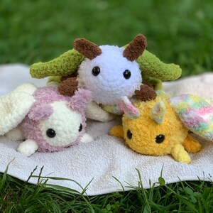 May include: Three crocheted stuffed animals with wings. The first is white with brown accents and green wings. The second is pink with white accents and white wings. The third is yellow with pink and blue accents and pink and blue wings.