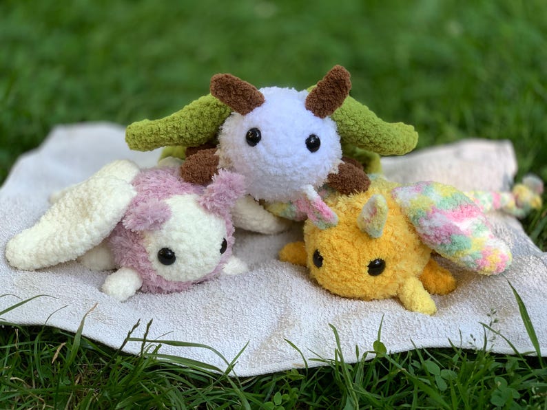 May include: Three crocheted stuffed animals with wings. The first is white with brown accents and green wings. The second is pink with white accents and white wings. The third is yellow with pink and blue accents and pink and blue wings.