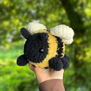 May include: A crocheted plush bee toy with black and yellow stripes, white wings, and black eyes. The bee is being held in a hand.