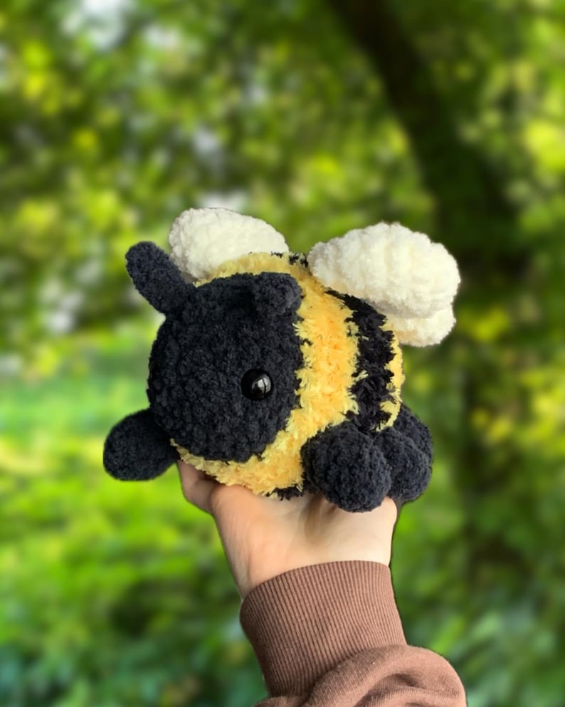 May include: A crocheted plush bee toy with black and yellow stripes, white wings, and black eyes. The bee is being held in a hand.
