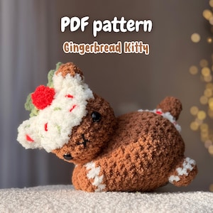 May include: A crocheted gingerbread kitty with white frosting, a red cherry, and green leaves. The kitty is brown with white accents and black eyes. The text "PDF pattern Gingerbread Kitty" is displayed above the kitty. The Instagram handle "@fiya.crochet" is at the bottom.