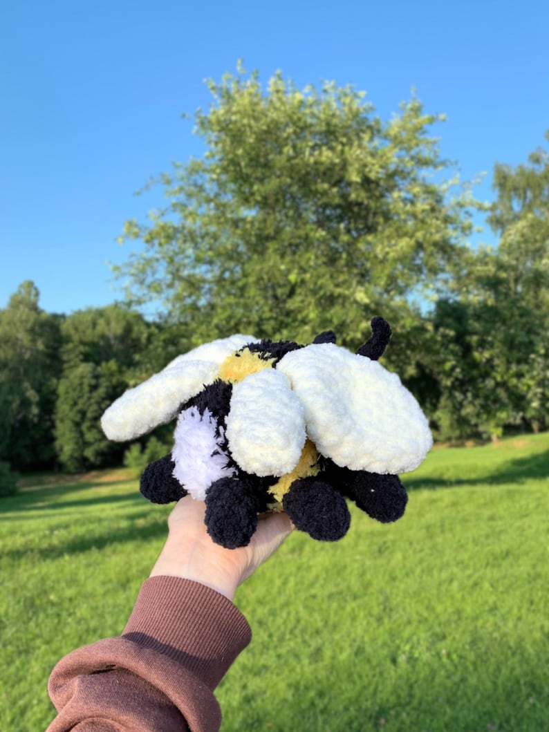 May include: A crocheted plush bee toy with black, white, and yellow yarn. The bee has a fuzzy texture and is holding its wings out.
