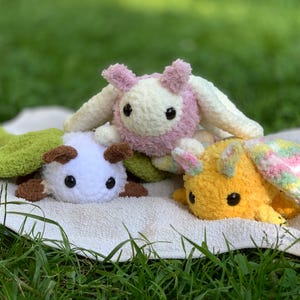 May include: Three crocheted stuffed animals, one pink and white, one green and white, and one yellow and white, are lying on a white towel. The animals are all facing the camera and have large, black eyes.