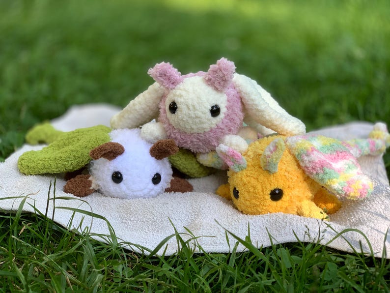 May include: Three crocheted stuffed animals, one pink and white, one green and white, and one yellow and white, are lying on a white towel. The animals are all facing the camera and have large, black eyes.
