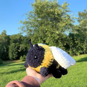 May include: A crocheted bumble bee plush toy with black and yellow fur, white wings, and black eyes. The toy is being held in a hand against a green grassy background.