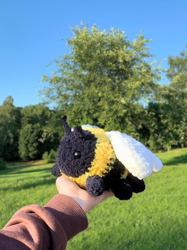 May include: A crocheted bumble bee plush toy with black and yellow fur, white wings, and black eyes. The toy is being held in a hand against a green grassy background.