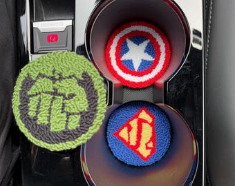 May include: Three round crocheted coasters featuring superhero logos. The coasters are green with a fist, red, white, and blue with a star, and blue with a yellow Superman logo. The coasters are in a car's cup holder.