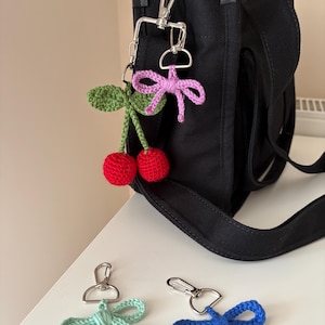 May include: A black tote bag with a silver clasp holding a crochet cherry charm with red cherries and green leaves, and a pink bow. Two additional crochet bow charms in blue and light green are also present.