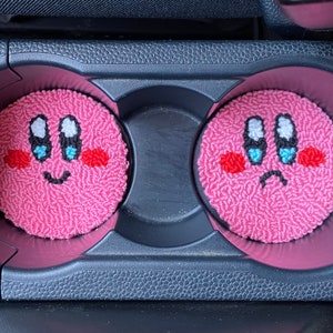 Peut inclure: Deux porte-gobelets de voiture roses avec des visages brodés d'un personnage rose aux yeux bleus et à la bouche blanche. Un visage sourit et l'autre fait la moue.