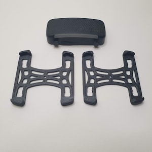 Puede incluir: Un conjunto de tres componentes negros impresos en 3D. La pieza superior es rectangular con una parte superior curvada y una superficie texturizada. Debajo hay dos soportes a juego con marco abierto. Los componentes son probablemente para un dispositivo.