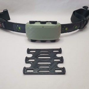 Puede incluir: Un collar de perro verde y negro con un pequeño dispositivo rectangular adjunto. El dispositivo tiene la palabra "Tractive" impresa en él. También se incluye un soporte de plástico negro.