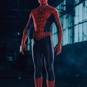 May include: A full-body shot of a person in a Spider-Man costume. The costume is red with a black spider emblem on the chest and a web pattern. The mask covers the entire head, and the background is a dark industrial setting.