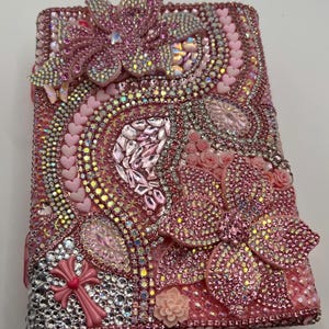May include: A rectangular notebook cover heavily embellished with pink and clear rhinestones, featuring large 3D flower appliqués, small pink heart-shaped beads, and a pink cross charm. The surface is densely covered in sparkling crystals.