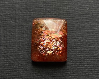 May include: A rectangular, iridescent sunstone cabochon with a reddish-brown base and a pattern of gold and silver flecks. The stone is cut in a faceted style.