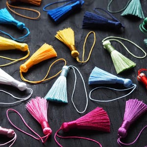 May include: A collection of multi-coloured silk-like tassels, each featuring a looped cord for hanging. The assortment includes vibrant shades of yellow, blue, pink, white, green, and red, scattered across the frame in various orientations.