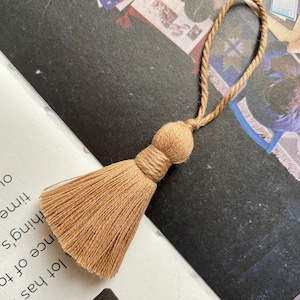 May include: A light brown tassel with a loop of twine. The tassel is made of soft, fluffy fibres and has a knotted top.