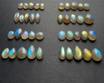 Puede incluir: Una colección de piedras preciosas de labradorita iridiscente en forma de lágrima en varios tonos de azul, verde y gris. Las piedras están dispuestas en filas sobre una superficie gris oscuro.