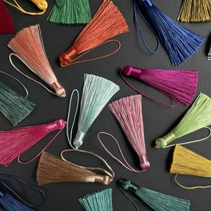 Puede incluir: Una colección de borlas decorativas con aspecto de seda en una variedad de colores vibrantes que incluyen rojo, azul, verde, rosa, naranja, plata y oro. Cada borla presenta un denso haz de hilos finos y un lazo a juego para colgar.