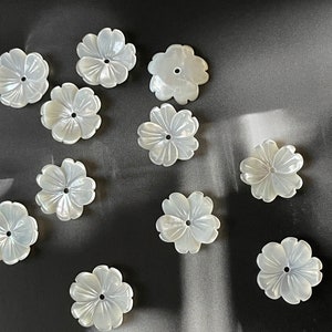 Puede incluir: Una colección de once cuentas blancas en forma de flor, con un brillo nacarado. Cada cuenta tiene cinco pétalos y un pequeño agujero en el centro. Las cuentas están dispersas sobre una superficie gris oscuro, con iluminación suave.