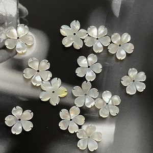 May include: A selection of iridescent, white flower-shaped beads. Each bead features five petals and a small central hole. The beads are arranged on a dark, textured surface, creating a delicate and elegant appearance.