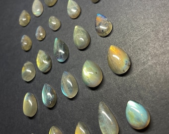 May include: A collection of 20 teardrop-shaped labradorite gemstones in various shades of gray, green, and blue. The stones are arranged in a staggered pattern on a black surface.