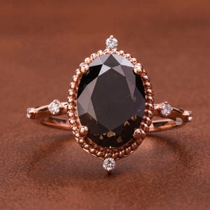 May include: Rose gold ring featuring a large, oval-cut, dark gemstone. The ring has a detailed, rope-like border around the center stone and smaller accent stones on the band. The ring is set against a brown background.