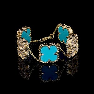 Könnte beinhalten: Ein goldfarbenes Armband mit türkisfarbenen Kleeblatt-Akzenten. Das Armband hat eine filigrane Kette und aufwendige Details um jedes Kleeblatt. Der Hintergrund ist schwarz, was das Schmuckdesign hervorhebt.