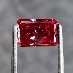 Könnte beinhalten: Ein rechteckiger facettierter Edelstein in tiefroter Farbe mit hoher Reinheit. Der Stein weist einen Brillantschliff mit zahlreichen dreieckigen und trapezförmigen Facetten auf, die das Licht reflektieren, und wird von zwei silberfarbenen Metallklemmen gehalten.