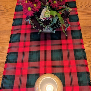 May include: A red and green plaid table runner with a fringed edge. The runner is decorated with a floral arrangement of burgundy flowers and greenery, and two candle holders with lit candles.