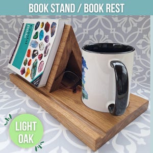 Puede incluir: Un soporte para libros de madera de roble claro. El soporte tiene forma triangular y puede sostener un libro y una taza. El soporte está sobre una superficie con un patrón.