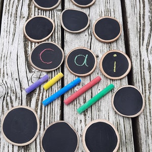 May include: A set of 10 round wooden chalkboard discs with black surfaces and natural wood edges. The discs are arranged in a semi-circle with four smaller discs in the centre. The smaller discs have the letters 'c', 'a', and 'i' written on them in different coloured chalk. Four pieces of chalk in different colours are also included.
