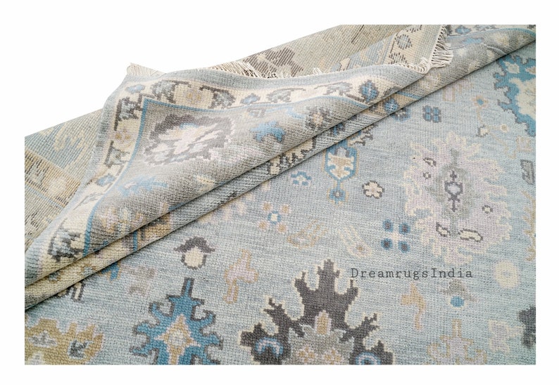 May include: A light blue rug with a traditional Persian design. The rug has a faded look and features intricate patterns in shades of blue, beige, and gray. The rug is folded in half, revealing the fringe on the edges. The text "DreamrugsIndia" is visible in the bottom right corner.